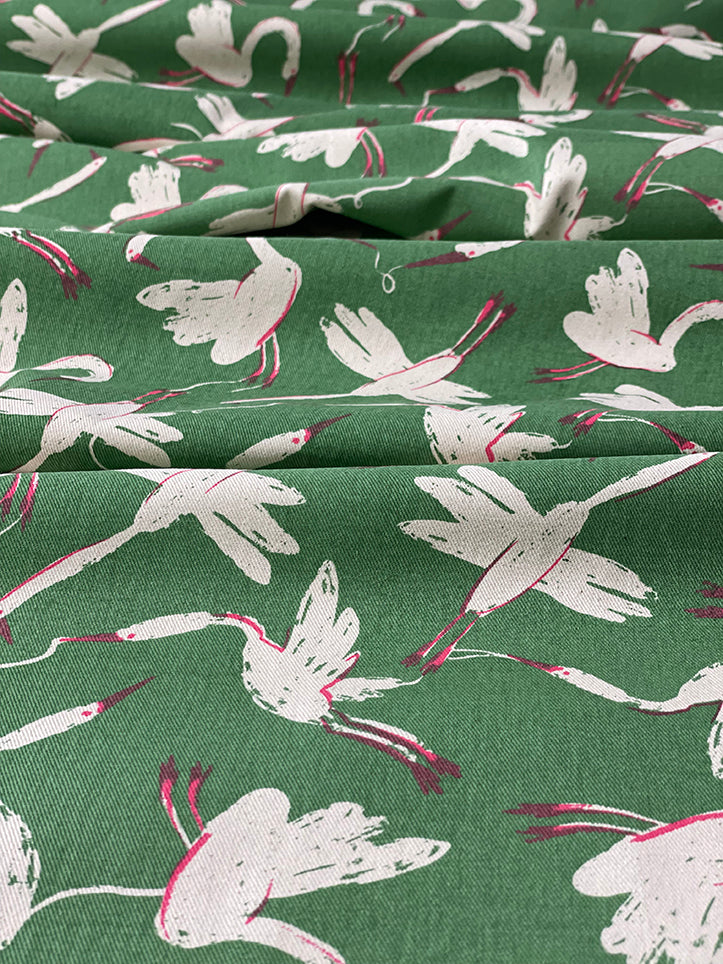 Fabric showing swan print. Vanilla base cloth. Printed green background around flying swans, with accents of hot pink on legs, beaks and shadows.